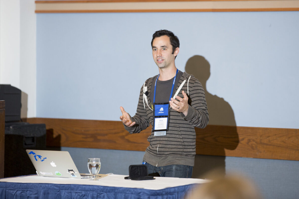 nick denardis speaking at a conference with laptop in front of him, hands outstretched